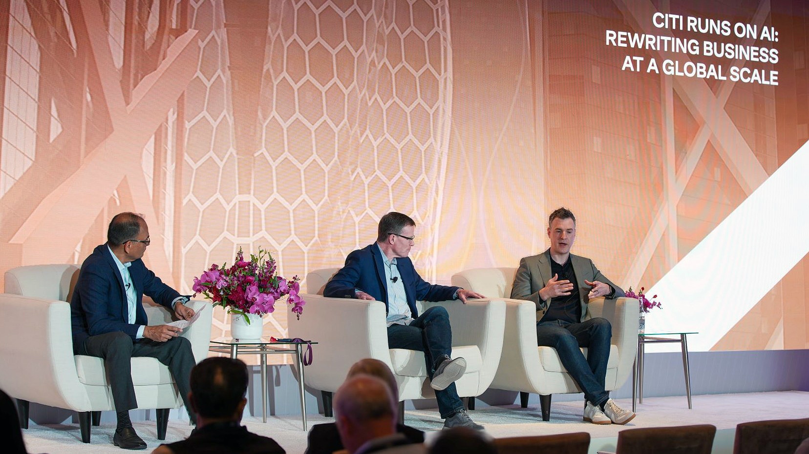This image captures a panel discussion on stage at a professional event. The moderator, Tim Ryan and David Griffiths are seated, engaged in conversation. The large background screen includes text that says, 