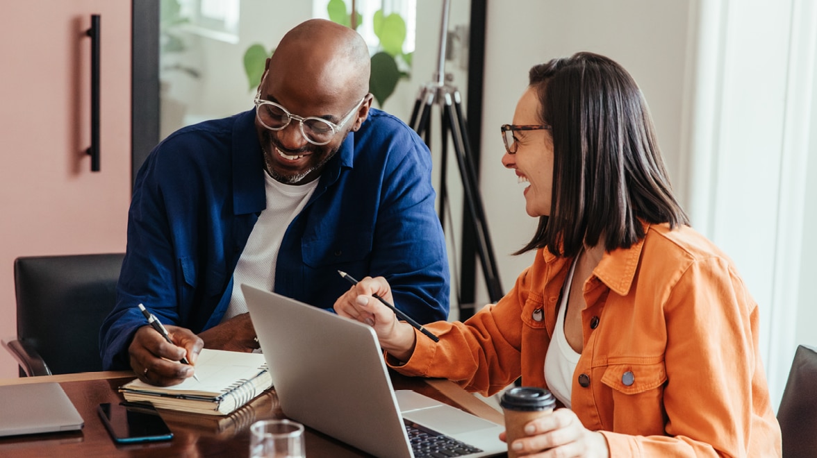 Two people laughing sitting at a table with a laptop and taking notes.
