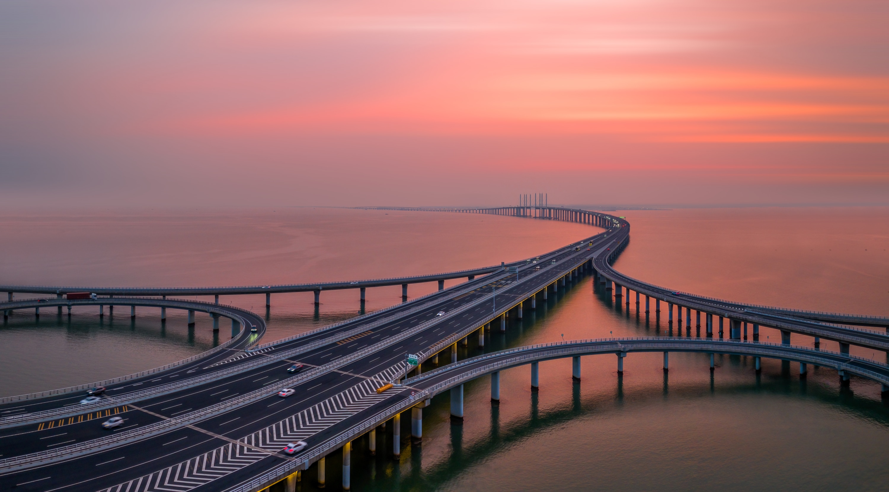 Bridge roads with cars over water with pink sunset in the background.