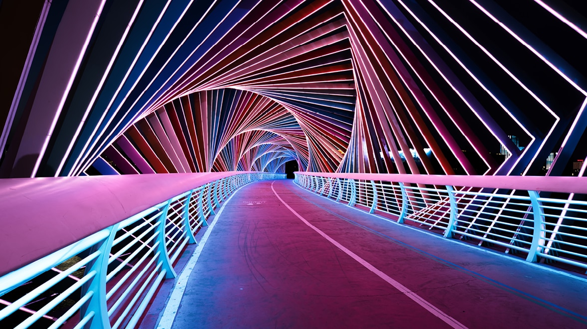 Modern pedestrian bridge illuminated with pink, blue and white lights, featuring repeating angular arches over a curved pathway.