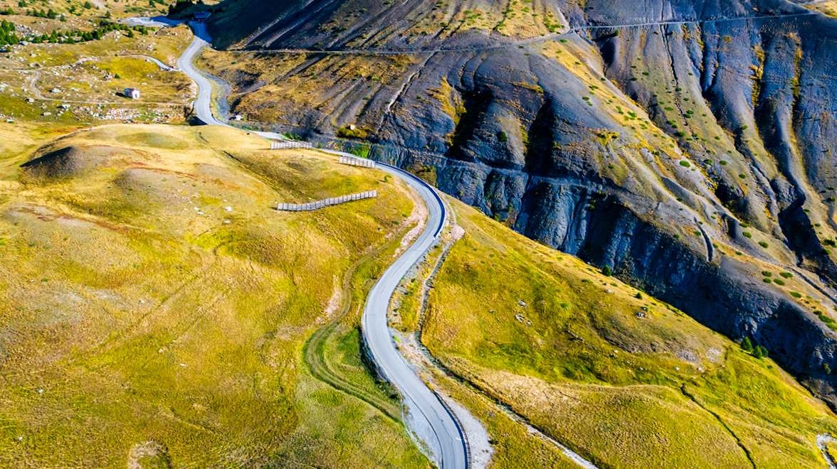 A winding paved road curves through grassy hills alongside steep, dark rocky mountain slopes.