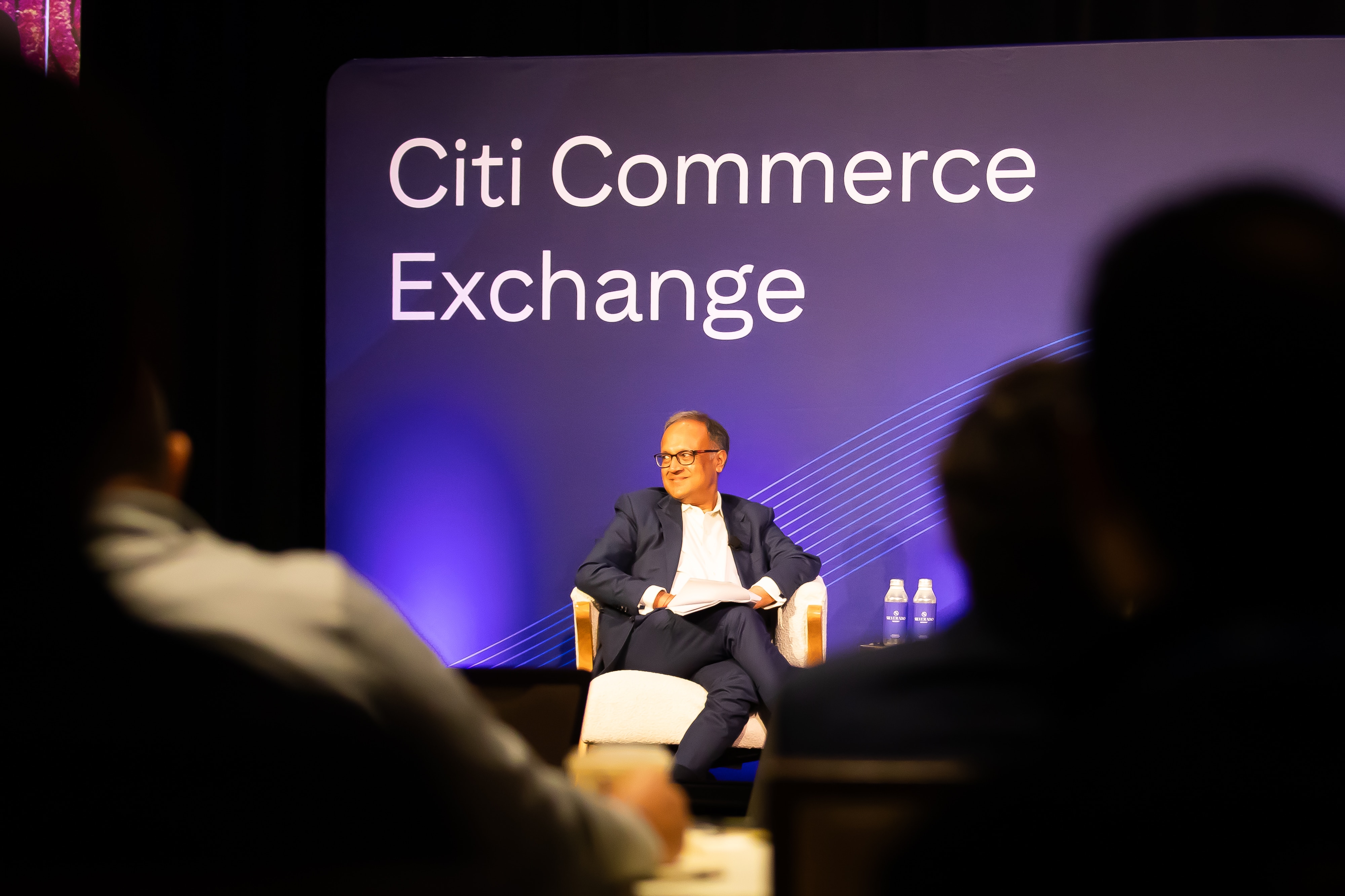 Ashish Bajaj, Head of Citi Services for North America, seated on stage during a session at the Citi Commerce Exchange event, viewed from the perspective of the audience. He's seated in front of a backdrop reading "Citi Commerce Exchange."